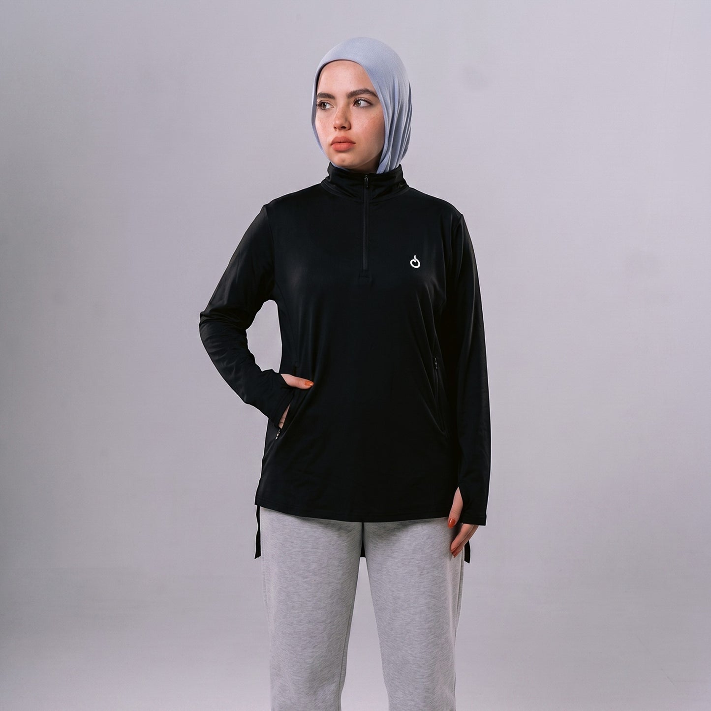 A model wearing a black Meryia WASEA’ Zip Top with a half-zip neckline and light blue hijab against a grey background.