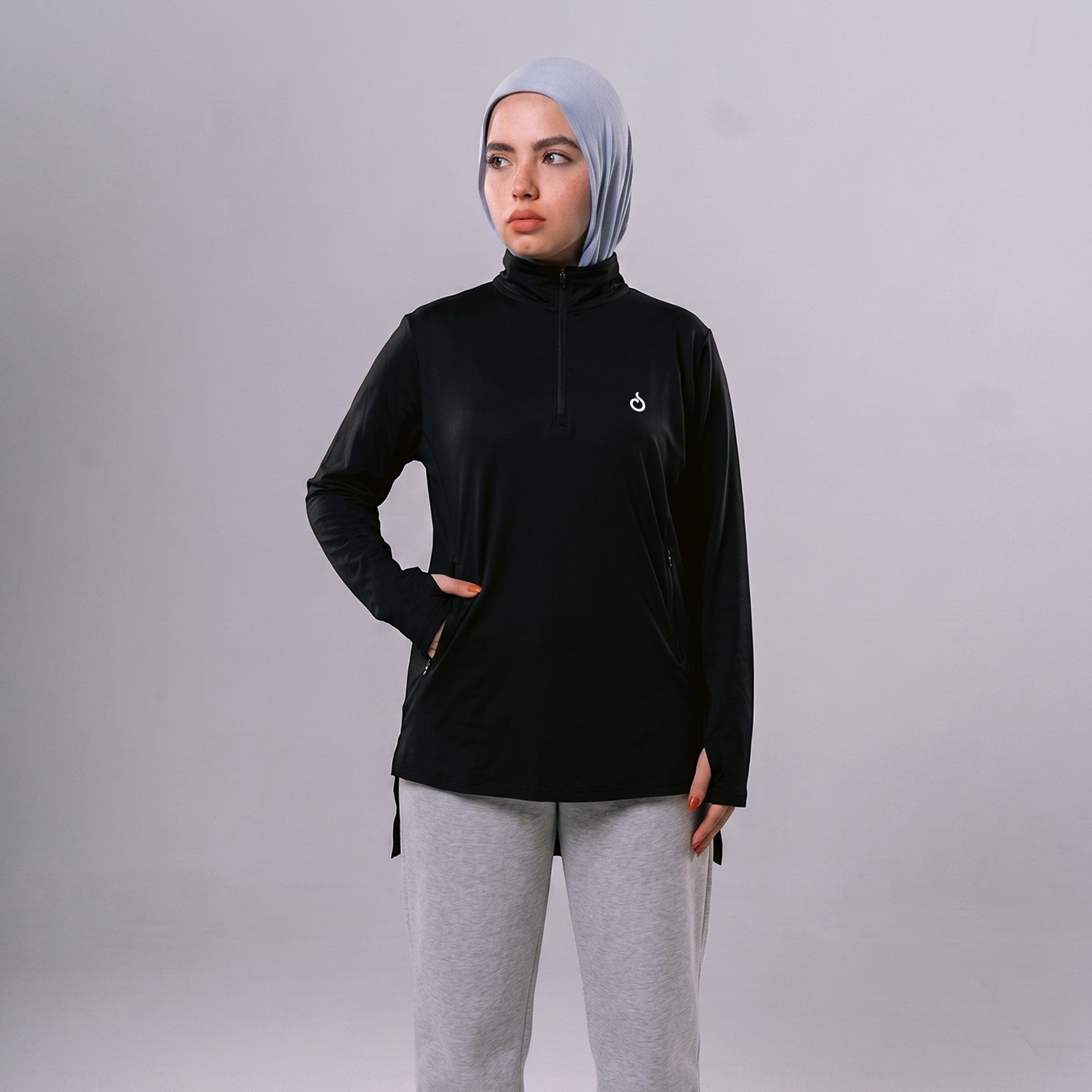 A model wearing a black Meryia WASEA’ Zip Top with a half-zip neckline and light blue hijab against a grey background.